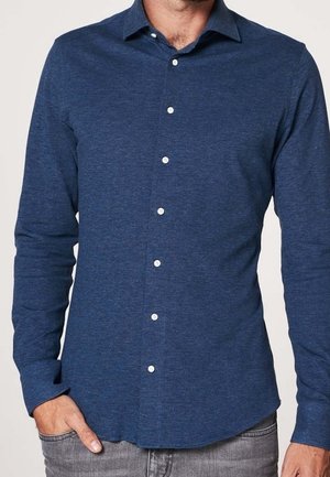 Man draagt een donkerblauw, lang mouws overhemd met knopen en een kraag, gecombineerd met lichtgrijze spijkerbroek, hand in zak, tegen een effen achtergrond.