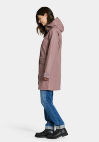 Vattentät rosa regnjacka med luva, sidfickor och justerbara muddar, kombinerad med blå jeans och svarta gummistövlar.