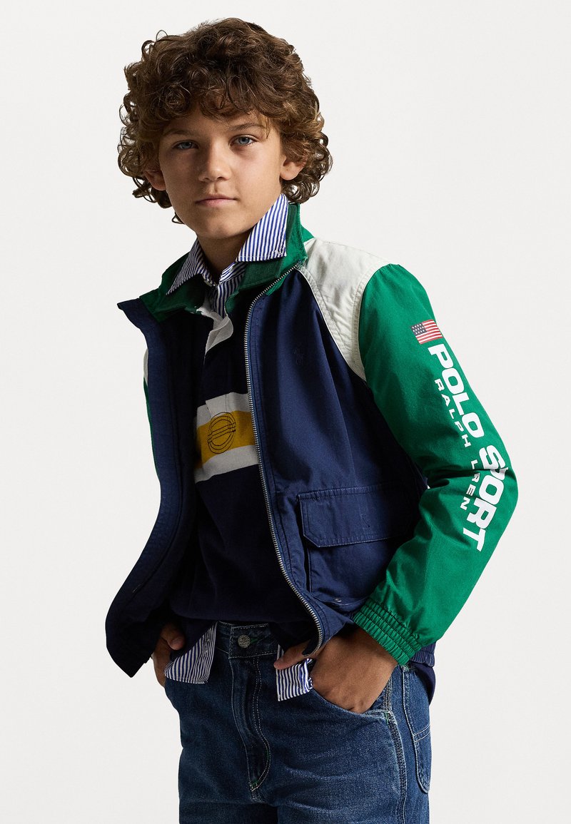 Χρωματιστό μπουφάν με πράσινα, μπλε και λευκά πάνελ; έχει την ένδειξη "Polo Sport" στη μανσέτα. Συνδυασμένο με πουκάμισο με ρίγες και τζην παντελόνι.