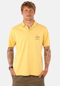 Polo shirt jaune en coton avec un col, des manches courtes et un logo sur le devant. Texture lisse et coupe décontractée.