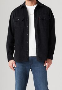 Camicia di cotone a coste nera con chiusura a bottoni, due tasche sul petto e collare. Indossata sopra una maglietta bianca e abbinata a jeans blu.