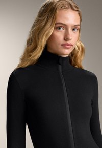 Top negru cu mâneci lungi, guler înalt și fermoar central. Confecționat dintr-un material neted și elastic, cu un design mulat.