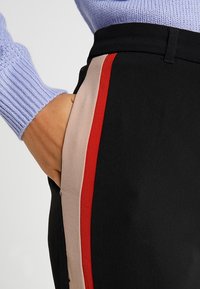 Ręka w kieszeni czarnych spodni z beżowymi i czerwonymi paskami po bokach, zestawionych z jasnofioletowym swetrem z prążków.