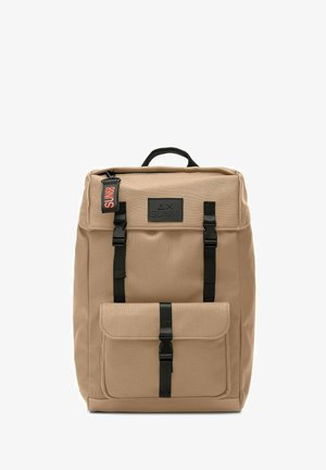 Sac à dos en toile beige avec des sangles et des éléments en métal noirs, doté d'une poche avant et d'une poignée supérieure. Étiquette logo remarquable en orange et noir.