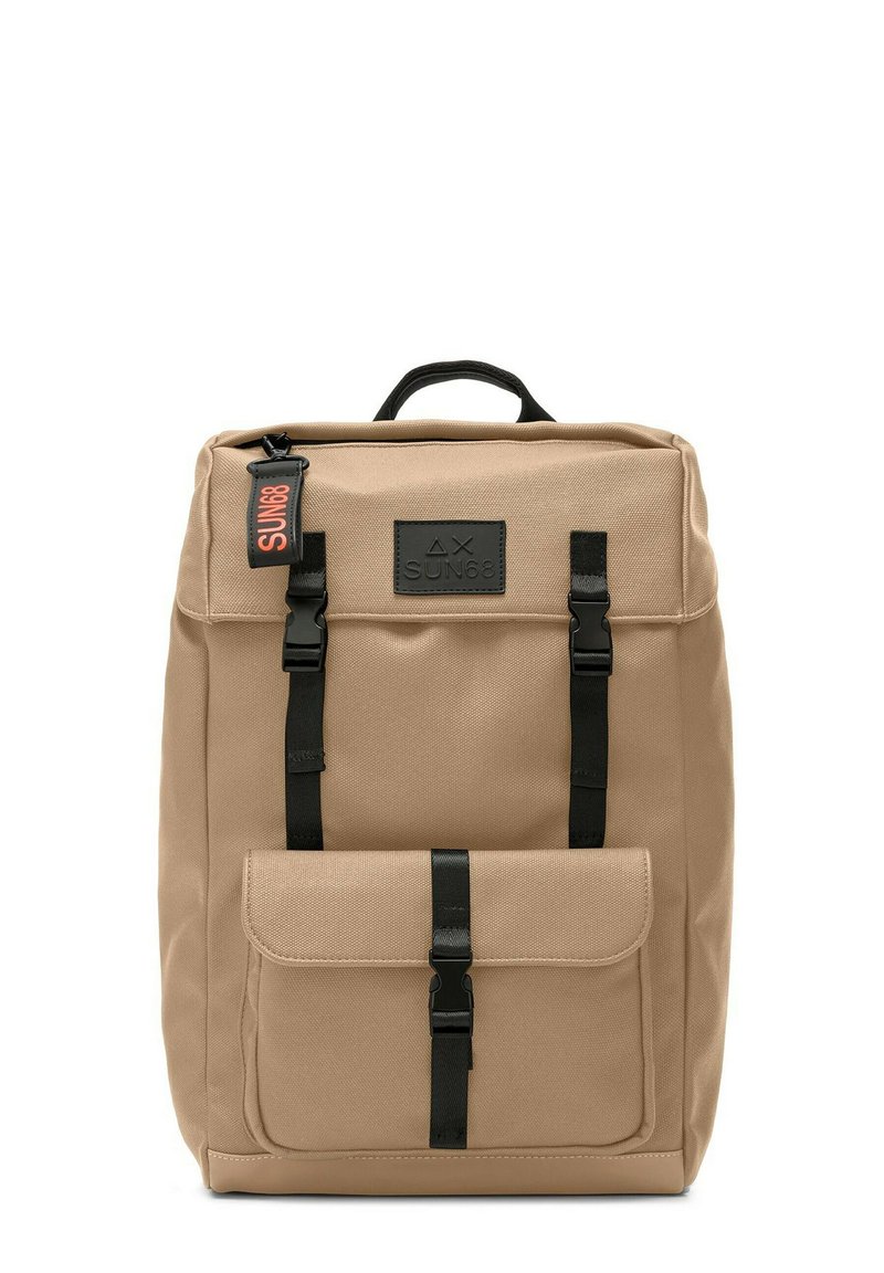 Sac à dos en toile beige avec des sangles et des éléments en métal noirs, doté d'une poche avant et d'une poignée supérieure. Étiquette logo remarquable en orange et noir.