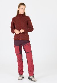 Röd, texturerad fleecjacka med hög krage och dragkedjefickor, kombinerad med röda och vinröda byxor som har cargofickor och dragkedjor.