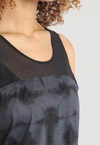 Kvinna med lockigt hår som bär en ärmlös svart och mörkgrå batiktop med en genomskinlig svart övre panel.