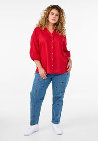 Röd skjorta med V-ringning och uppvikta ärmar, kombinerad med blå jeans som har röd läppbroderi och vita sneakers.