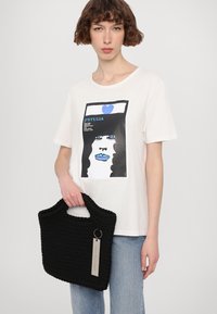 Schwarze Strickhandtasche mit grober Textur und einem oberen Griff, kombiniert mit einem weißen T-Shirt mit einem grafischen Druck eines Frauenansichts in Blau und Schwarz.