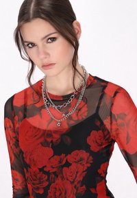 Durchsichtige schwarze Bluse mit rotem Blumenmuster, Rundhalsausschnitt, geschichtet mit mehreren silbernen Ketten und einem herzförmigen Anhänger, was einen strukturierten Look erzeugt.