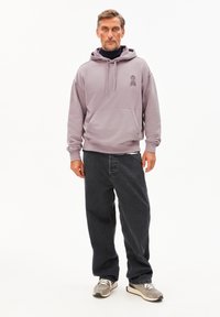 Uomo che indossa un hoodie viola chiaro sopra un dolcevita nero, jeans neri larghi e sneakers grigie, in piedi contro uno sfondo bianco semplice.