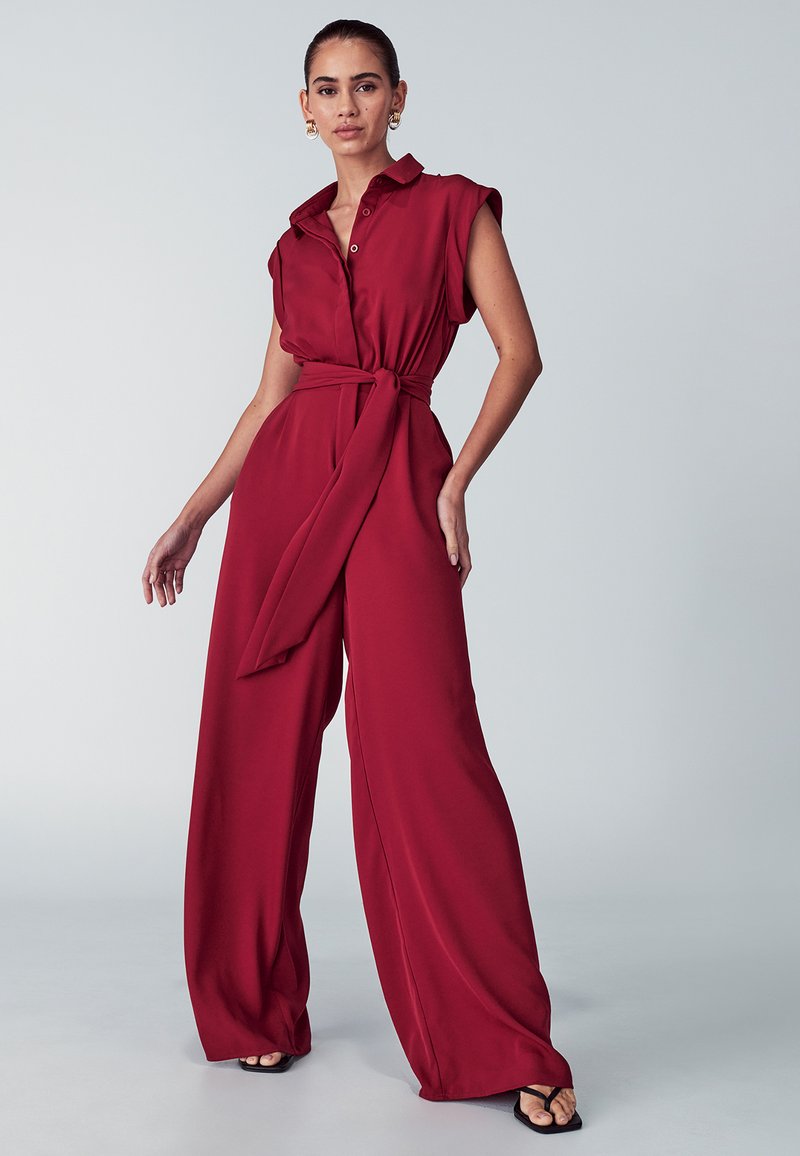 Rdeč kombinezon s ovratnikom, kratkimi rokavi in zavezanem pasu. Ima široke noge in gladko tkanino.