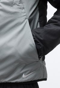 Άτομο ντυμένο με μπουφάν Nike δίχρωμο σε γκρι και μαύρο, με το χέρι στην πλαϊνή τσέπη, που δείχνει την υφή του υφάσματος και το λογότυπο Nike κοντά στο τελείωμα.