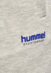 Hellgrauer strukturierter Stoff mit blauem "hummel" und grauem "SPORTSWEAR" Logo, auf den Brustbereich der Kleidung gestickt.