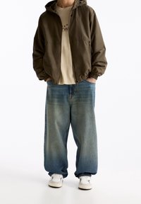 Chaqueta con capucha de color verde oliva con puños elásticos, camiseta beige y jeans de mezclilla azul claro sueltos. Zapatillas blancas completan el conjunto.