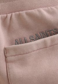 Lys rosa stof med ribbet tekstur og en lille lomme. "ALLSAINTS" broderet i grå på lommen. Simpel design, blødt materiale.