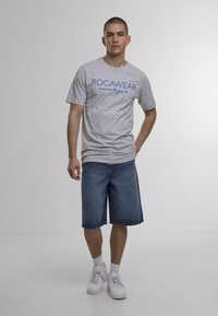 Grå bomulds t-shirt med blå "ROCAWEAR" tekst, parret med denim shorts til knæene. Modellen bærer hvide sneakers og ankelstrømper.
