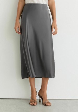 Kvinde iført en grå satin midi nederdel med høj talje og en let sideslids, kombineret med beige stroppede højhælede sandaler.