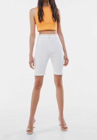 Bermudas de mezclilla blanca con puños enrollados, combinadas con una camiseta de tirantes naranja de canalé. El conjunto presenta un diseño ajustado y una cintura alta.