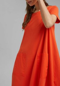 Kvinna iförd en löst sittande, kortärmad klänning i ljus orange färg med två stora framfickor, stående mot en plain grå bakgrund.