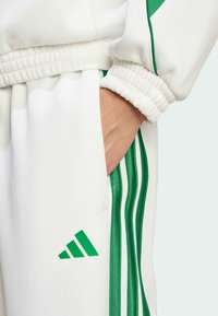 Balts sporta tracksuits ar zaļu trīs svītru dizainu. Izceļas ar mīkstu tekstūru, elastīgām aprocēm un zaļu Adidas logo uz kājas.