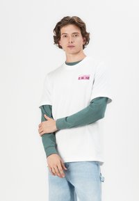 Hvit t-skjorte med korte ermer lagt over en grønn langermet skjorte. Har et rosa "adidas Originals" trykk på brystet. Lys blå jeans.