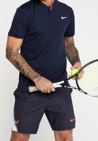 Marinblå pikétröja med henley-krage och vit Nike-logotyp, kombinerad med mörka shorts med orange detalj och ett tennisracket.