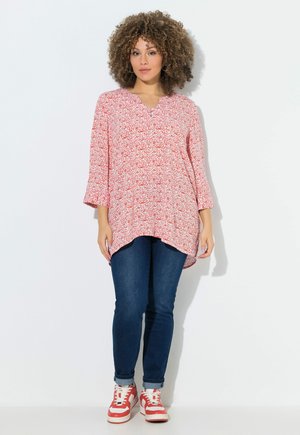 Vrouw met krullend haar, gekleed in een rode tuniek met patroon, donkerblauwe jeans en rood-witte sneakers, staand tegen een witte achtergrond.