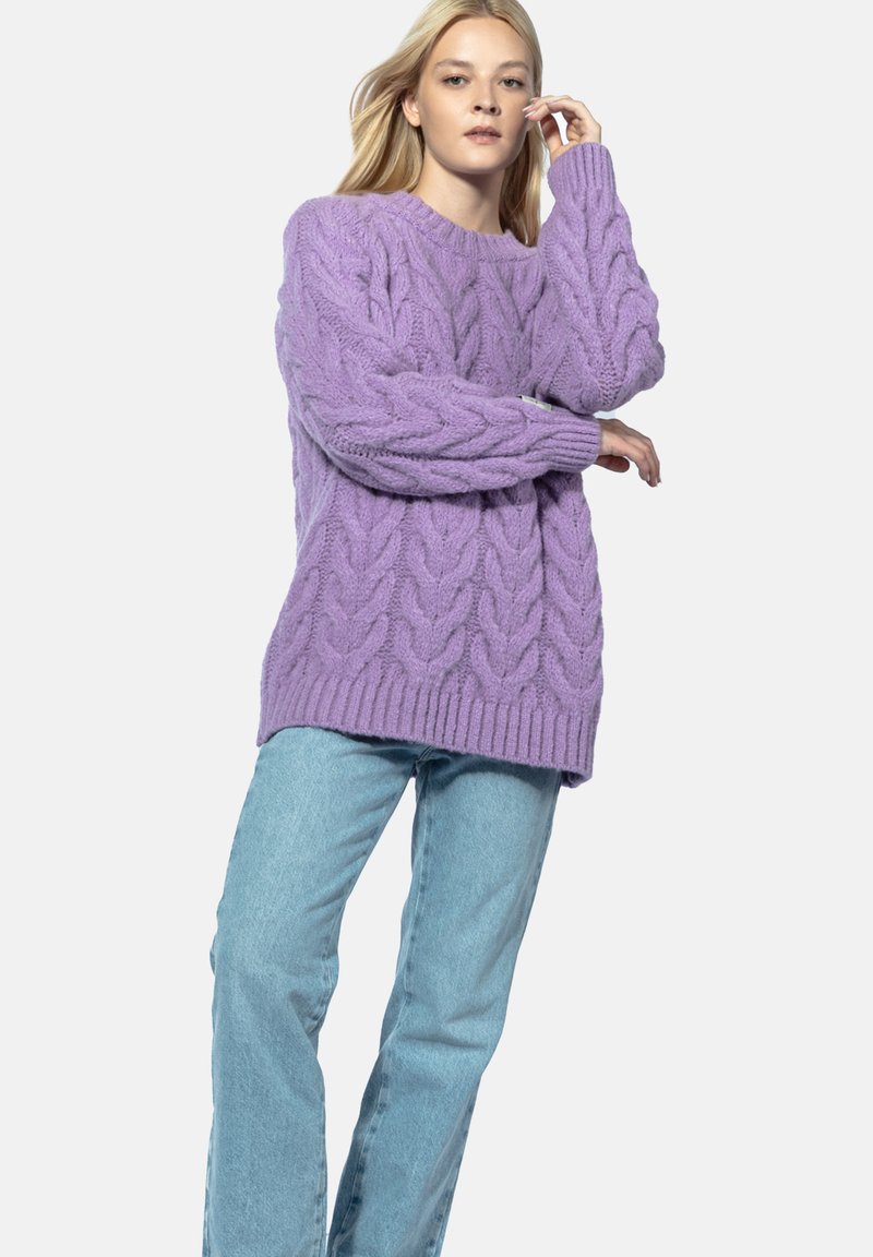Sweter w warkocze w kolorze lawendowym, charakteryzujący się luźnym krojem, ściągaczami u rękawów i u dołu oraz grubą teksturą przędzy. Połączony z jasnoniebieskimi dżinsami.