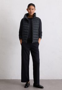Femme se tenant debout avec les mains dans les poches, portant un gilet rembourré noir sur une chemise à manches longues, un pantalon noir et des mocassins noirs devant un mur uni.