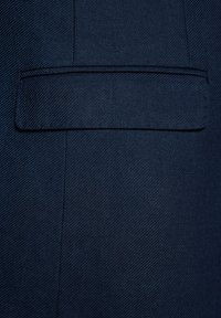 Niebieski żakiet z tkaniny w drobnym diagonalnym prążku. Posiada jedną płaską kieszeń po lewej stronie oraz bezszwowe przeszycia wzdłuż krawędzi.