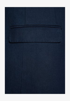 Niebieski żakiet z tkaniny w drobnym diagonalnym prążku. Posiada jedną płaską kieszeń po lewej stronie oraz bezszwowe przeszycia wzdłuż krawędzi.