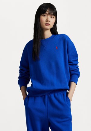 Blue sweatshirt made of soft fabric, featuring a crew neck, ribbed cuffs, and a small red logo on the chest, paired with matching pants.