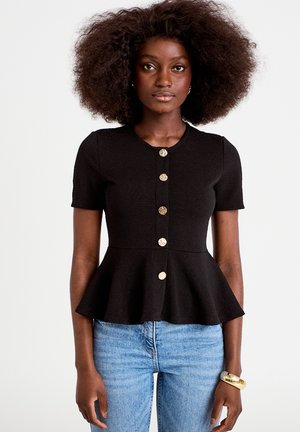 Kobieta z bujnymi kręconymi włosami ma na sobie czarny krótki top z basenem i złotymi guzikami oraz jasnoniebieskie dżinsy na jednolitym tle.