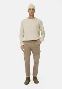 Beige langærmet skjorte, lysebrune corduroy cargo bukser med sidelommer og olivengrønne snørebåndsstøvler. En strikket cremefarvet hue på hovedet.