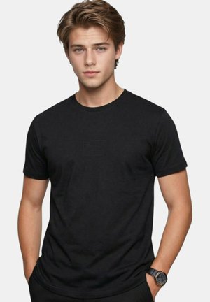 Jeune homme aux cheveux châtain clair portant un T-shirt noir uni Lee Cooper et une montre-bracelet noire, debout devant un fond blanc uni.