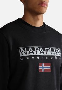 Homme portant un sweat-shirt noir avec le logo "NAPAPIJRI geographic" et un petit drapeau norvégien sous le texte sur la poitrine.