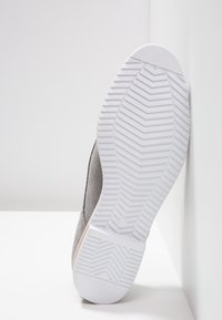 Semelle de chaussure avec un motif à chevrons blanc, inclinée contre un mur et un sol blancs, montrant la surface supérieure grise texturée.