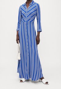 Niebieska i biała maxi sukienka w wzory z kołnierzykiem, długimi rękawami i zwiewnym krojem. Para białych sandałów na słupku z odkrytymi palcami.