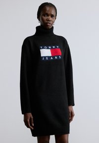 Musta neulemekko, jossa korkea kaulus ja rohkea kirjailtu "TOMMY JEANS" -logo sekä väriplokki-muotoilu punaisessa, valkoisessa ja sinisessä.