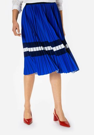 Kobieta nosząca plisowaną niebieską spódnicę w czarno-białe paski, zestawioną z czerwonymi sandałkami na obcasie typu slingback.