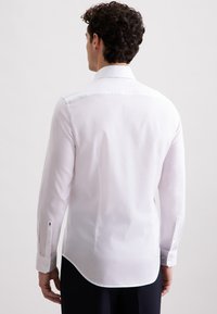 Uomo dai capelli ricci, che indossa una camicia bianca a maniche lunghe e pantaloni scuri, in piedi di spalle contro uno sfondo neutro.