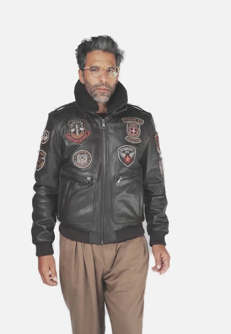 Homme aux cheveux et à la barbe grise, portant des lunettes transparentes, une veste en cuir noire avec des patchs de style militaire et un pantalon marron ample.