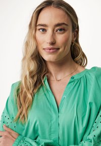 Blusa verde a bottoni con maniche lunghe, caratterizzata da intagli ricamati. Texture liscia e vestibilità ampia, abbinata a una delicata collana dorata.