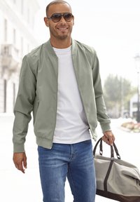 Hellgrüne Bomberjacke, weißes T-Shirt, blaue Jeans. Hält eine graue Sporttasche mit dunkelbraunen Akzenten. Sonnenbrille rundet den Look ab.