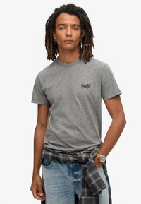 Grijs T-shirt met korte mouwen en een geribbelde halslijn, met een zwart logo op de borst. Gecombineerd met versleten blauwe spijkerbroeken en een geruite blouse die om de taille is geknoopt.