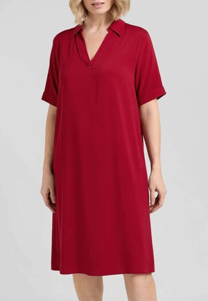 Donna che indossa un vestito rosso a maniche corte, lungo fino al ginocchio, con colletto e scollo a V, su sfondo grigio uniforme.