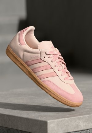 Lichtroze Adidas Samba sneaker met gumzool en drie donkerder roze strepen, getoond zwevend boven een grijze betonnen ondergrond.