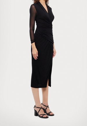 Vrouw draagt een zwart midi-jurkje met doorschijnende lange mouwen en een geplooide taille, gecombineerd met zwarte sandalen met bandjes en blokhakken.