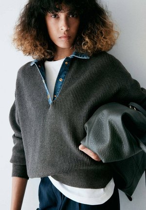Una modelo con cabello rizado lleva un suéter gris acanalado sobre una camisa vaquera y sostiene un gran bolso de cuero negro.